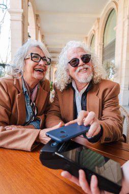 Keyifli yaşlı çift, neşeli bir kafede ödeme yapmak için akıllı telefonlarını kullanıyor. Kahkahalarla dolu özel bir anı paylaştıklarında neşe ve bağlantı yayıyorlar.