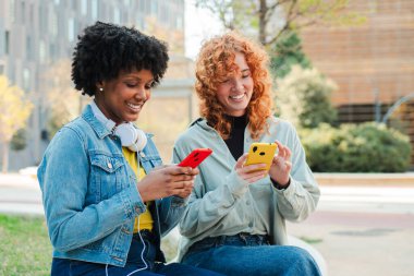 İki neşeli arkadaş gülümserken akıllı telefonlarıyla etkileşim kurarak birlikte vakit geçiriyorlar. Mobil cihazlarına odaklanırlar, anlarını ve deneyimlerini birbirleriyle paylaşırlar..