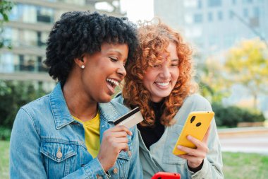 İki neşeli kadın, akıllı telefonları ve kredi kartlarıyla birlikte alışverişin neşesini canlı bir şekilde sergiliyorlar..