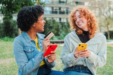 İki neşeli kadın aktif olarak bir cep telefonu ödeme sürecine katılıyor, gülümsüyor ve akıllı telefonları ve bir kredi kartıyla meşgul oluyorlar, online alışveriş ve ödeme basitliğinin heyecanını sergiliyorlar.