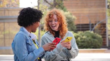 İki neşeli arkadaş gülümserken akıllı telefonlarıyla etkileşim kurarak birlikte vakit geçiriyorlar. Mobil cihazlarına odaklanırlar, anlarını ve deneyimlerini birbirleriyle paylaşırlar..