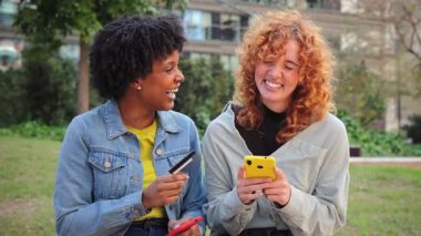 İki neşeli kadın, akıllı telefonları ve kredi kartlarıyla birlikte alışverişin neşesini canlı bir şekilde sergiliyorlar..