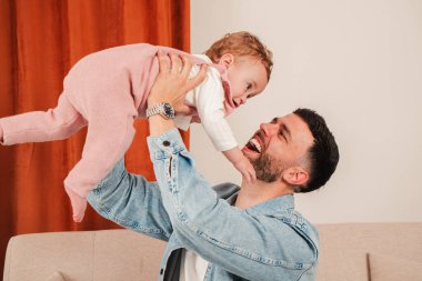 Neşeli bir baba, gülümseyen bebeğini havaya kaldırırken neşeli bir etkileşim içinde olur. İçi kahkaha ve sevgiyle dolu, aile ve ebeveynliğin özünü somutlaştıran bir an yaşar..