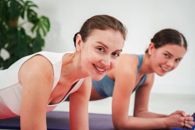 İki hevesli kadın birlikte tahta egzersizi yapıyor, kameraya gülümserken güç ve destek gösteriyorlar, takım çalışması ruhunu somutlaştırıyorlar ve neşeli bir atmosferde sağlıklı bir yaşam sürüyorlar..