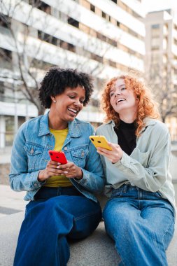 İki hayat dolu genç kadın, bu canlı ve anlamlı fotoğrafta neşeli bir şekilde akıllı telefonlarıyla gülüşlerini ve keyifli anlarını paylaşıyor, arkadaşlıklarını ve bağlantılarını sergiliyor..