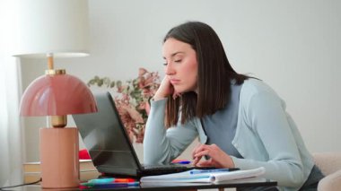 Genç bir kadın dizüstü bilgisayarıyla uğraşırken stres ve tefekkür belirtileri gösteriyor. Evde otururken akademik zorlukları ve öğrenci hayatının baskılarını yansıtıyor.