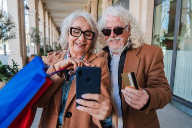 Joyful senior couple enjoying their special shopping day together, happily taking a selfie with a smartphone to capture their delightful moments and sharing laughter while holding shopping bags filled