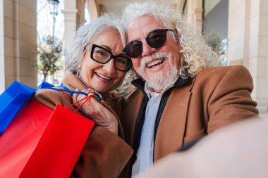 A lively senior couple enjoying their time together, taking a fun selfie while holding colorful shopping bags. They are filled with joy and warmth, showcasing their love and happiness during their