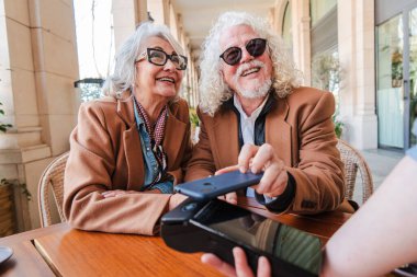 Delighted senior couple enjoying a moment of connection while making a mobile payment with their smartphone and credit card, showcasing a blend of technology and love in their lives as they bond