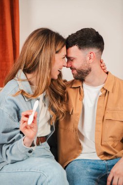 Happy couple sharing an intimate moment while joyfully holding a pregnancy test, radiating love and excitement in a cozy indoor atmosphere with warm colors and affectionate expressions.
