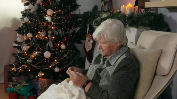 一名老年妇女编织一条毯子 坐在附近的圣诞树壁炉旁边的椅子上 — 图库视频影像