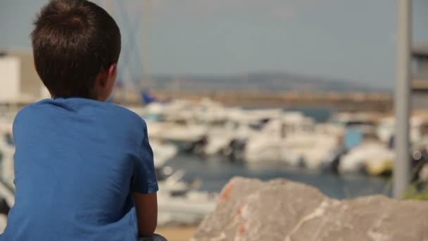 Jongen zit op steen en kijkt naar de baai met jachten — Stockvideo