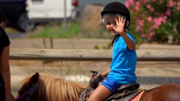 Petit garçon en t-shirt bleu monte un poney, agite sa main et montre un gros pouce — Video