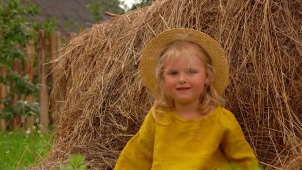 Söt liten flicka i en halmhatt sitter på en höstack — Stockvideo