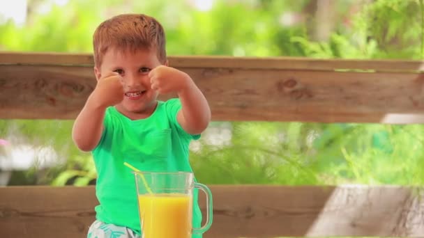 The cheerful little boy is showing big thumbs on the background of orange juice — Stock Video