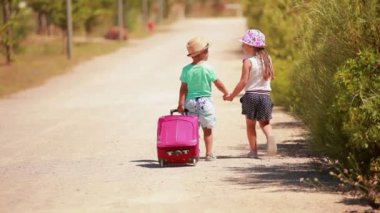 Şapkalı çocuklar ellerinde pembe bir bavulla yol boyunca el ele yürüyorlar.