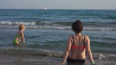 Annem deniz kıyısında oturuyor ve kızının deniz dalgalarıyla oynamasını izliyor.