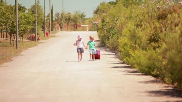 El niño y la niña en sombreros están caminando de la mano a lo largo de la carretera con una maleta — Vídeo de stock