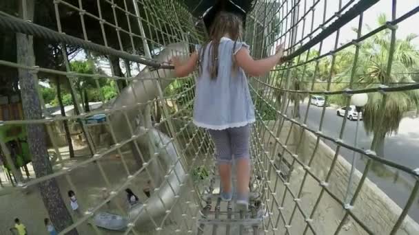 Вид сзади на маленькую девочку, идущую по высокому безопасному мосту на детской площадке — стоковое видео