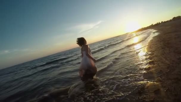 Μια γυναίκα με λευκό φόρεμα τρέχει κατά μήκος της παραλίας προς το ηλιοβασίλεμα — Αρχείο Βίντεο
