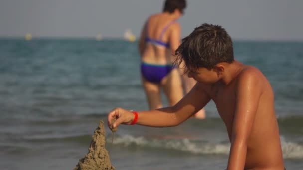 O rapaz bronzeado está a construir um castelo de areia. — Vídeo de Stock