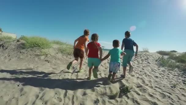 Vidám gyerekek fényes ruhában futkároznak a homokos tengerparton a tengerhez. — Stock videók