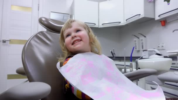 Ένα μικρό χαριτωμένο κορίτσι κάθεται σε μια οδοντιατρική καρέκλα και χαμογελά — Αρχείο Βίντεο
