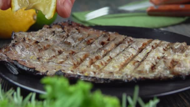 La mano está exprimiendo jugo de limón en el pescado blanco a la parrilla en el plato negro — Vídeos de Stock