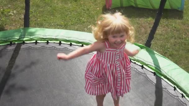 Una niña alegre en un vestido está saltando en el trampolín en el patio trasero — Vídeo de stock