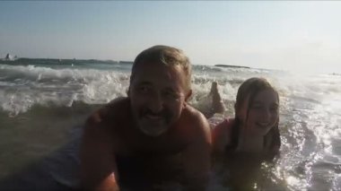 Baba ve kız su sıçratan deniz dalgalarında yüzükoyun yatar ve mutlu bir şekilde gülümserler.