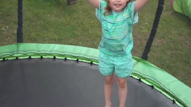 Küçük bir kız zıplıyor ve arka bahçede trambolinde el sallıyor. — Stok video