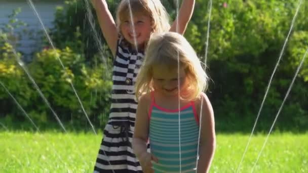 Dos niñas lindas están saltando alrededor de un aspersor de agua giratorio en el césped — Vídeo de stock