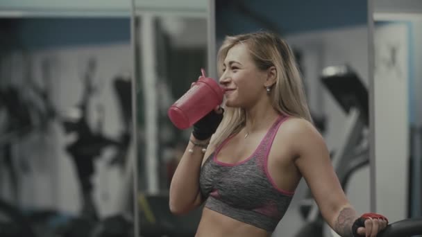 Hermosa chica bebiendo proteína en el gimnasio. La hembra bebe agua después del ejercicio — Vídeo de stock