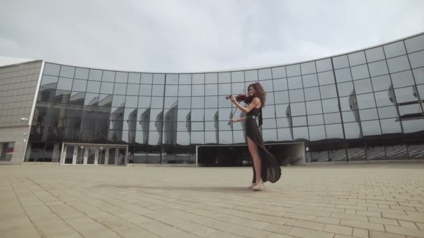 Schöne Frau in schwarzem Kleid spielt Geige in der Nähe von Glasgebäude. Kunstkonzept — Stockvideo