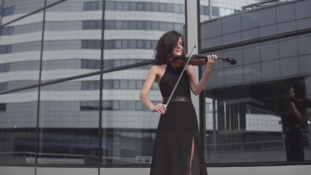 Елегантна жінка в чорній сукні грає на скрипці біля скляної будівлі. Художня концепція — стокове відео