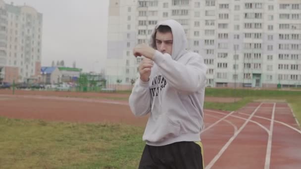 Young man training in hoodie boxing with invisible opponent at stadium. — Stock Video