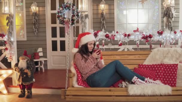 Ładna dziewczyna w Boże Narodzenie kapelusz picia herbaty, siedząc na ławce Choinka — Wideo stockowe