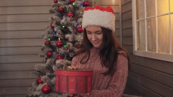 Chica joven con sombrero de Navidad regalo abierto. Chica sonríe con regalo de Navidad en la mano — Vídeos de Stock