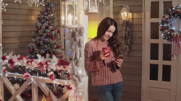 Mujer sonriente bebe té uso de teléfono en el porche con árbol de Navidad en el fondo — Vídeo de stock