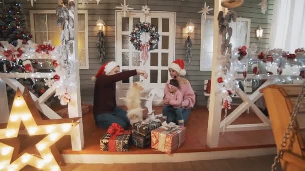 Lachende gezin met kleine dochter en hond zittend op de veranda met kerstboom — Stockvideo