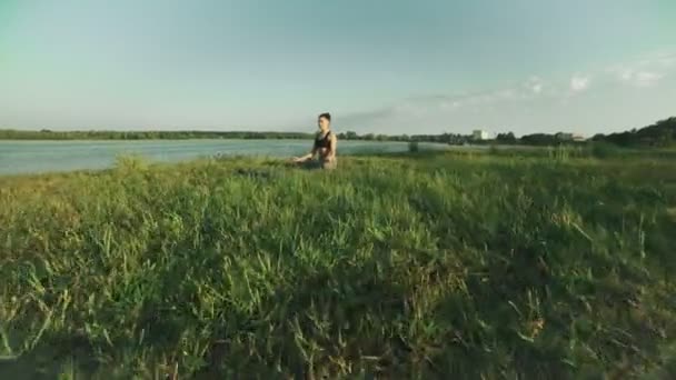 Jeune fille en position lotus au lever du soleil. Femme faisant du yoga le matin au lac — Video