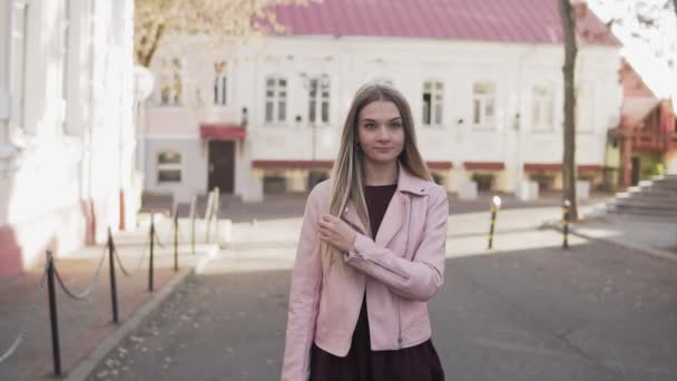 Красива дівчина в рожевій куртці ходить по місту і посміхається на камеру в повільному темпі — стокове відео