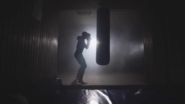 Боксер бьет грушу для битья в тёмном зале. Молодой человек тренируется в помещении — стоковое видео