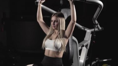 Blok Simulator koyu spor salonunda sarışın kız omuz üzerinde egzersiz yaparak kasları