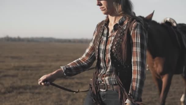 Cowgirl, die bei Sonnenaufgang ihr dunkles Pferd am Zügel führt. mächtiges Pferd zu Fuß — Stockvideo