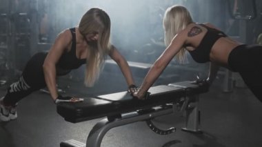 Şınav bankta gerçekleştirme iki Sportif sarışın kız. Fitness genç kadınlar gönderme yapıyor ups yavaş koyu spor salonunda. Orta atış