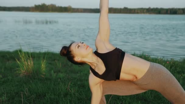 美丽的年轻女孩穿着运动服黑色上衣和灰色紧身衣做瑜伽在早晨在湖在蓝色瑜伽垫在安静的风景 做瑜伽的女人特写 — 图库视频影像