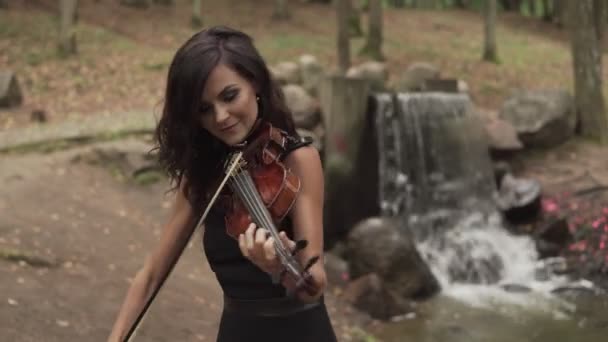 Schönes Mädchen in schwarzem Kleid spielt Geige im Wald mit Wasserfall — Stockvideo