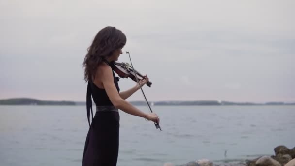 美丽的女孩在黑色礼服演奏小提琴在海背景。4k 中的艺术概念 — 图库视频影像