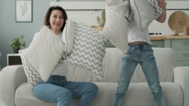 Anne ve küçük oğlu yastık kusuyor. Küçük oğlu oturma odasında kanepede anne ile komik yaşıyor. Erkek ve anne gömlek ve kot yavaş çekimde birlikte oynuyor